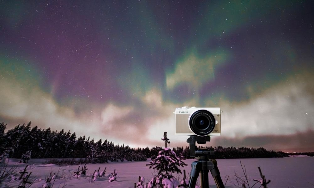 A travel camera on a tripod set up in a Finnish Lapland snow clearing photographing vivid green and violet Northern Lights above a silhouetted pine forest treeline