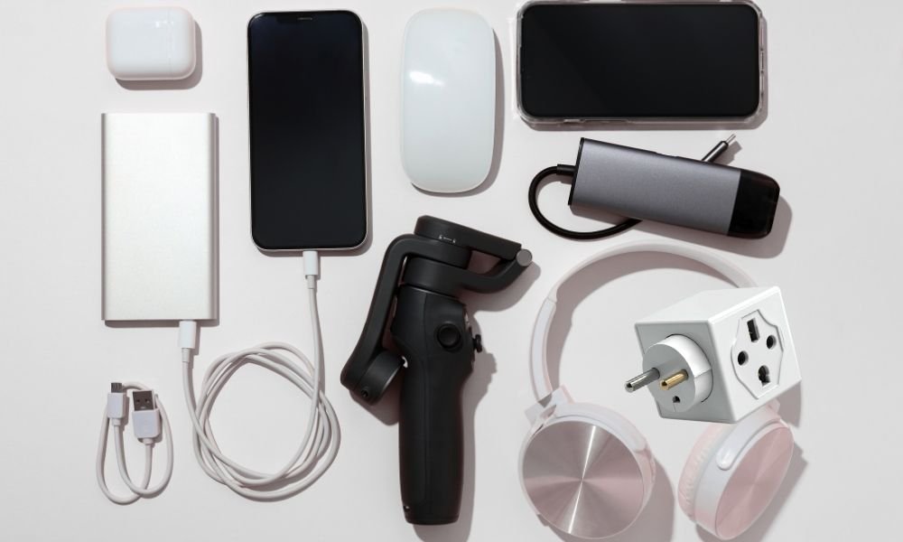 Flat lay of Scandinavia travel tech essentials including a power bank, universal travel adapter, wireless earphones, headlamp, and mirrorless camera on a dark slate surface