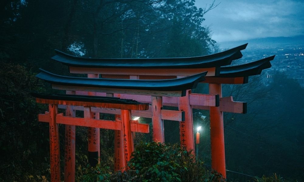 Empty torii gate path at Fushimi Inari in Kyoto at 6 AM, mist between orange gates, single hiker silhouette in distance, forest dark on both sides