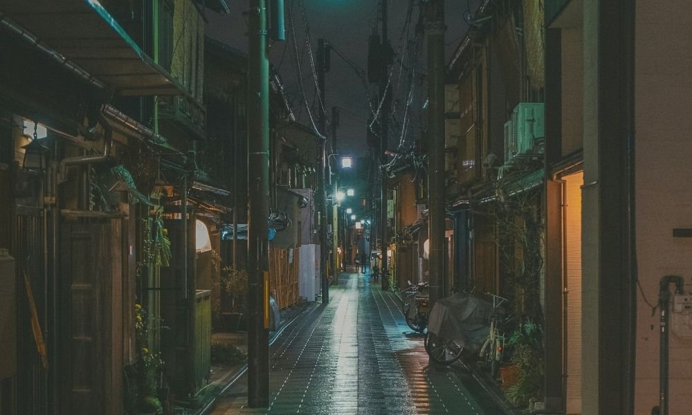 Solo woman walking through well-lit Gion street in Kyoto at night, lanterns reflecting on wet pavement, no crowds, peaceful atmosphere