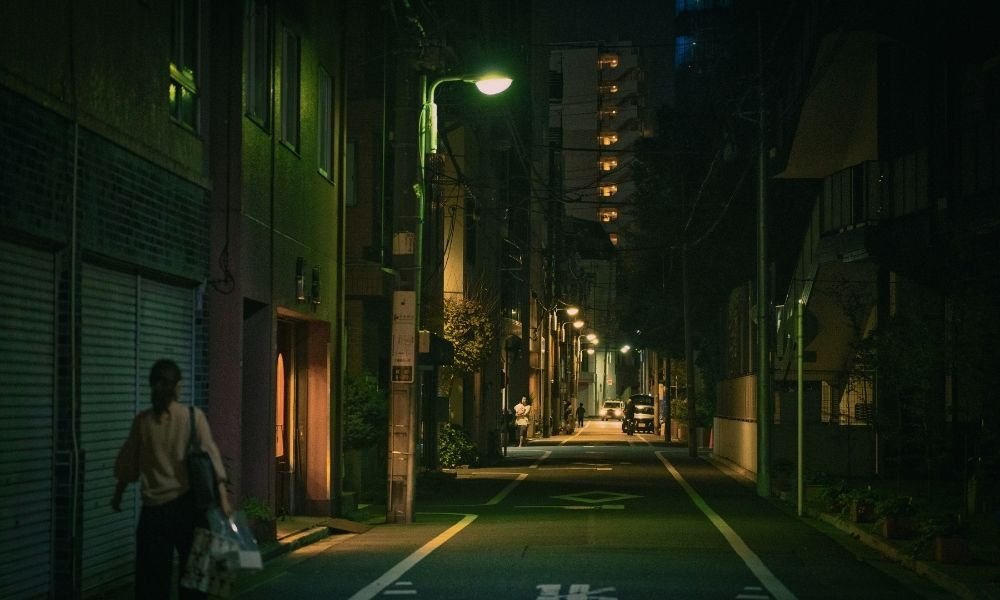 Well-lit Kyoto street at night, convenience store with bright signage, solo woman walking with bag, residential buildings, sense of safety