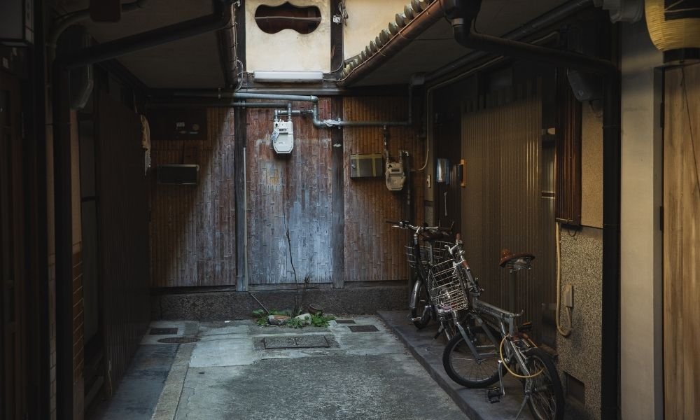 Narrow alley behind Nishiki Market in Kyoto, knife shop with blades in window, tofu maker's noren curtain, delivery bicycle parked, morning light