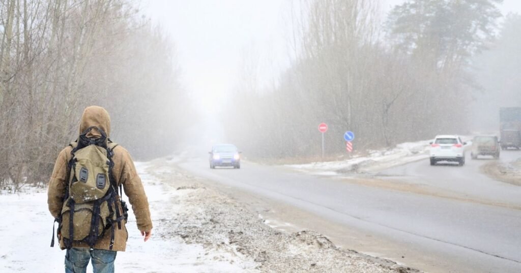 Solo traveler with backpack walking away down a misty dirt road in early morning light, long shadow stretching ahead