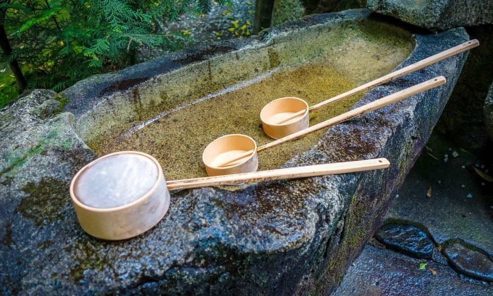 Water basin at Kyoto shrine entrance, wooden ladles arranged, hands being washed in ritual purification, stone lantern in background, morning light