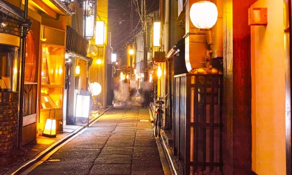 Narrow Pontocho Alley in Kyoto at night with lanterns, wooden restaurant facades, and the Kamogawa river visible through gaps