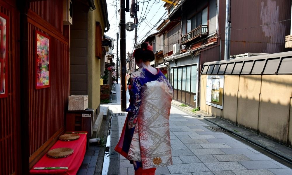 `Geiko walking through a quiet Gion side street in Kyoto, photographed from a respectful distance`