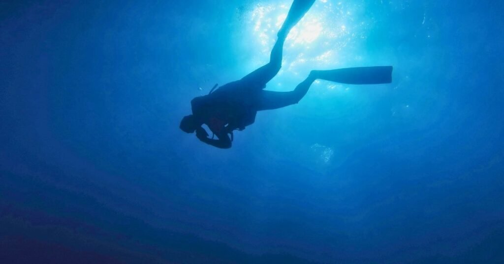 Scuba diver descending into deep blue water seen from above the surface, small against the vast dark below