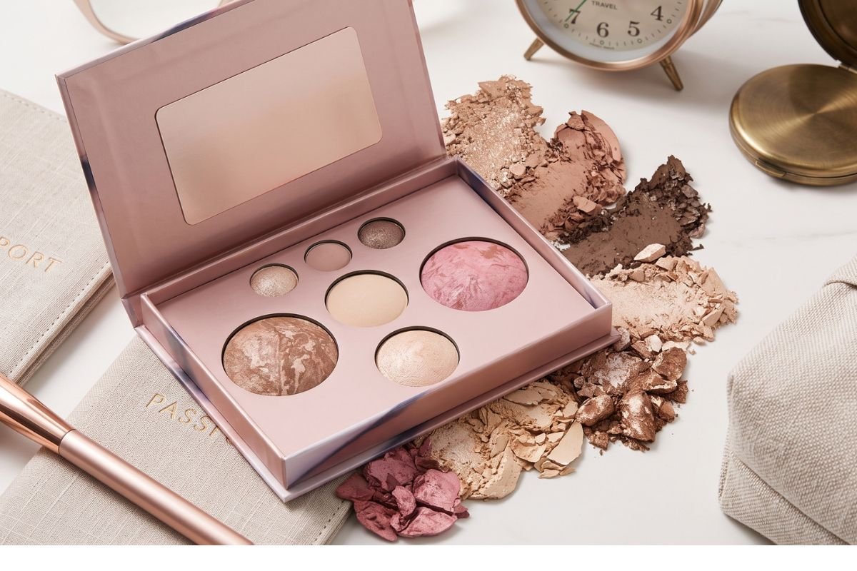 A pristine, opened rose-gold all-in-one makeup palette with nine different face and eye powders, centered on a light surface with scattered matching cosmetic powders and curated travel items like a brush and passport holder.