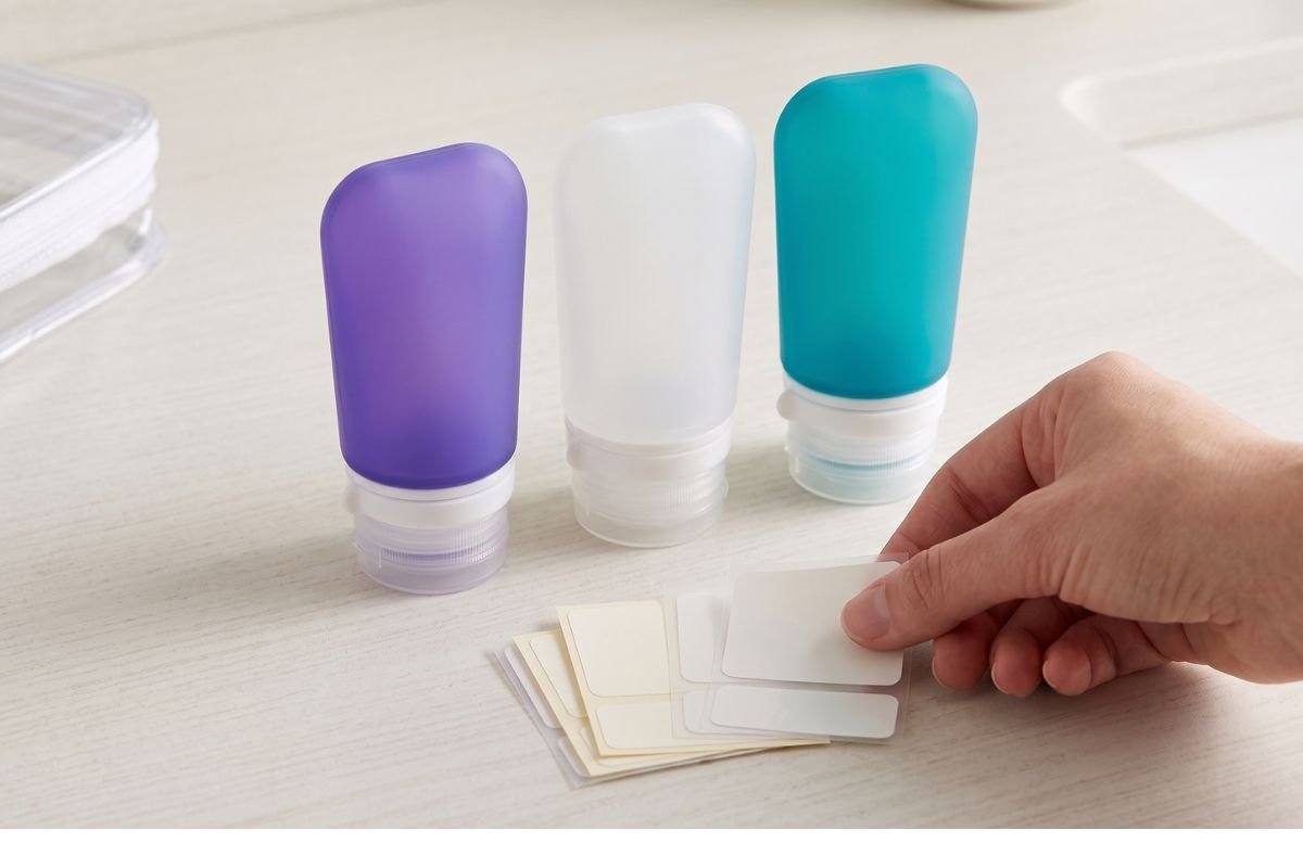 An artful composition of the three unbranded silicone travel bottles (purple, clear, teal) on a clean wooden surface, next to a stack of unprinted adhesive labels, on a light wood background with blurred context.