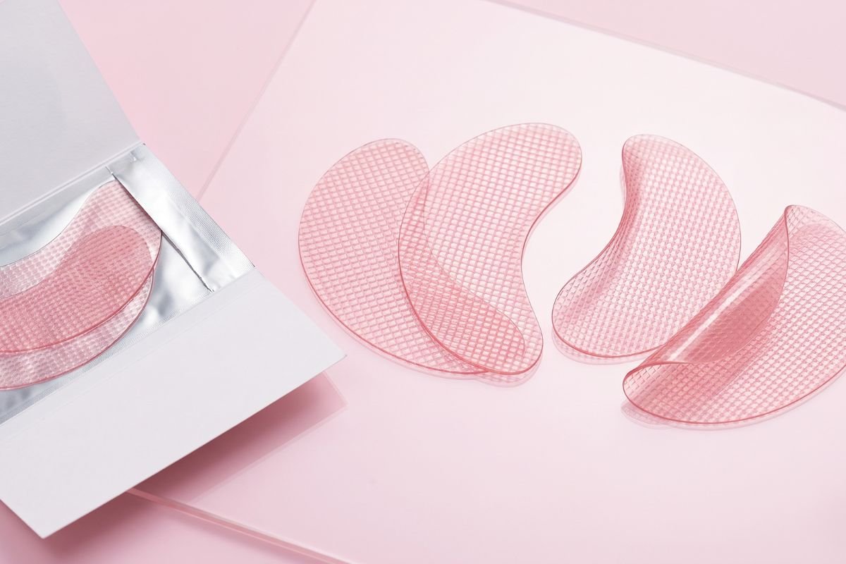 Three pairs of textured clear gel under-eye patches on a light pink surface, fanned out, next to an open paper envelope showing another pair.